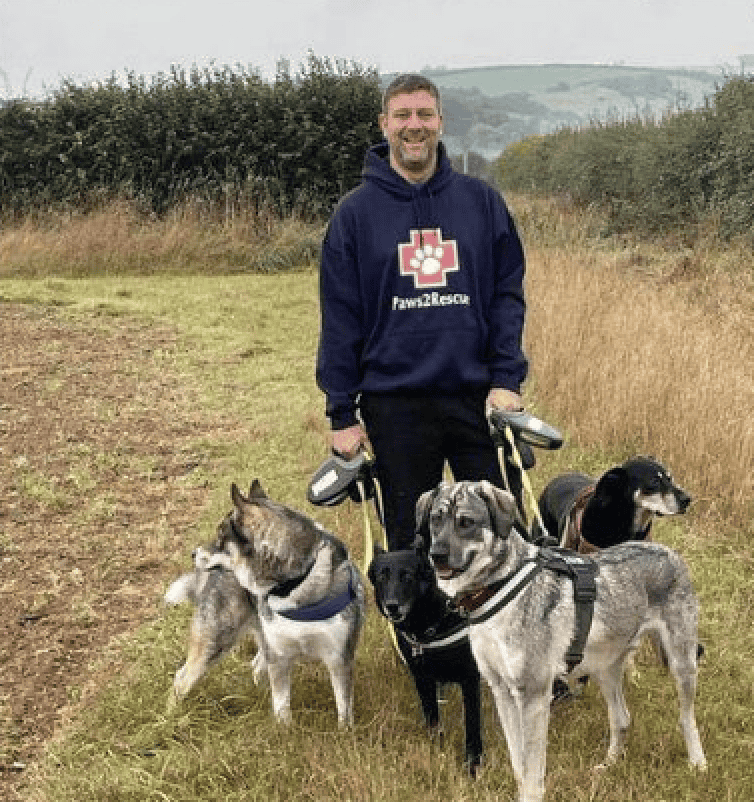 A photo of Paws2Rescue team member Darren standing in a field wearing a hoodie with the Paws2Rescue logo printed on the front, he is surrounded by a group of happy dogs